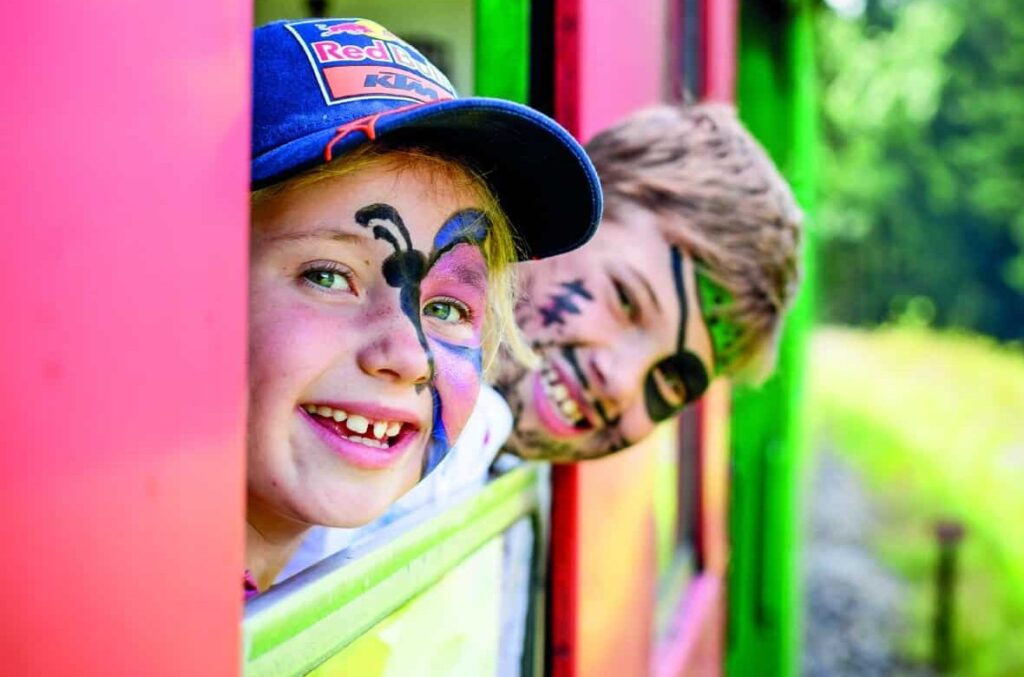 Zwei geschminkte Kinder schauen während einer Fahrt mit dem Murtalbahn-Kinderzug aus dem Waggon.