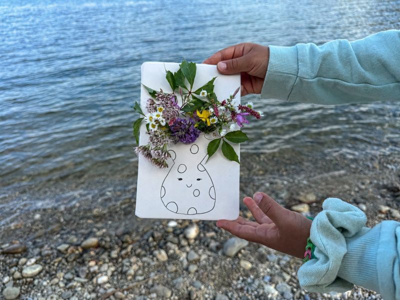 Kind präsentiert eine selbst gebastelte Naturvase mit bunten Wildblumen am See.